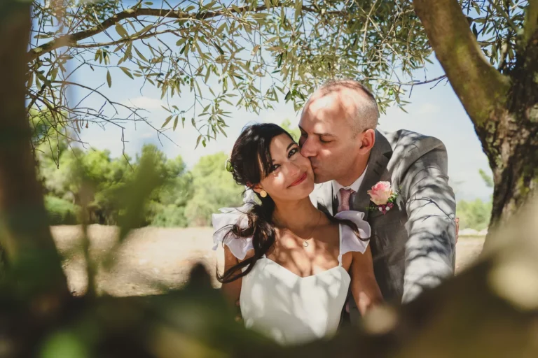 couple de mariés se faisant un bisou sur la joue dans un parc sous les oliviers pour leur mariage